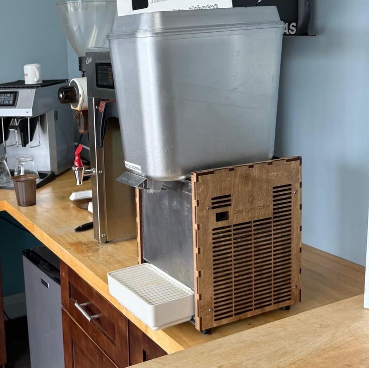 The original Cold Cycle Coffee prototype on a cafe counter: a bulky stainless mixing tank sitting on a rough laser-cut plywood box that houses the refrigeration unit
