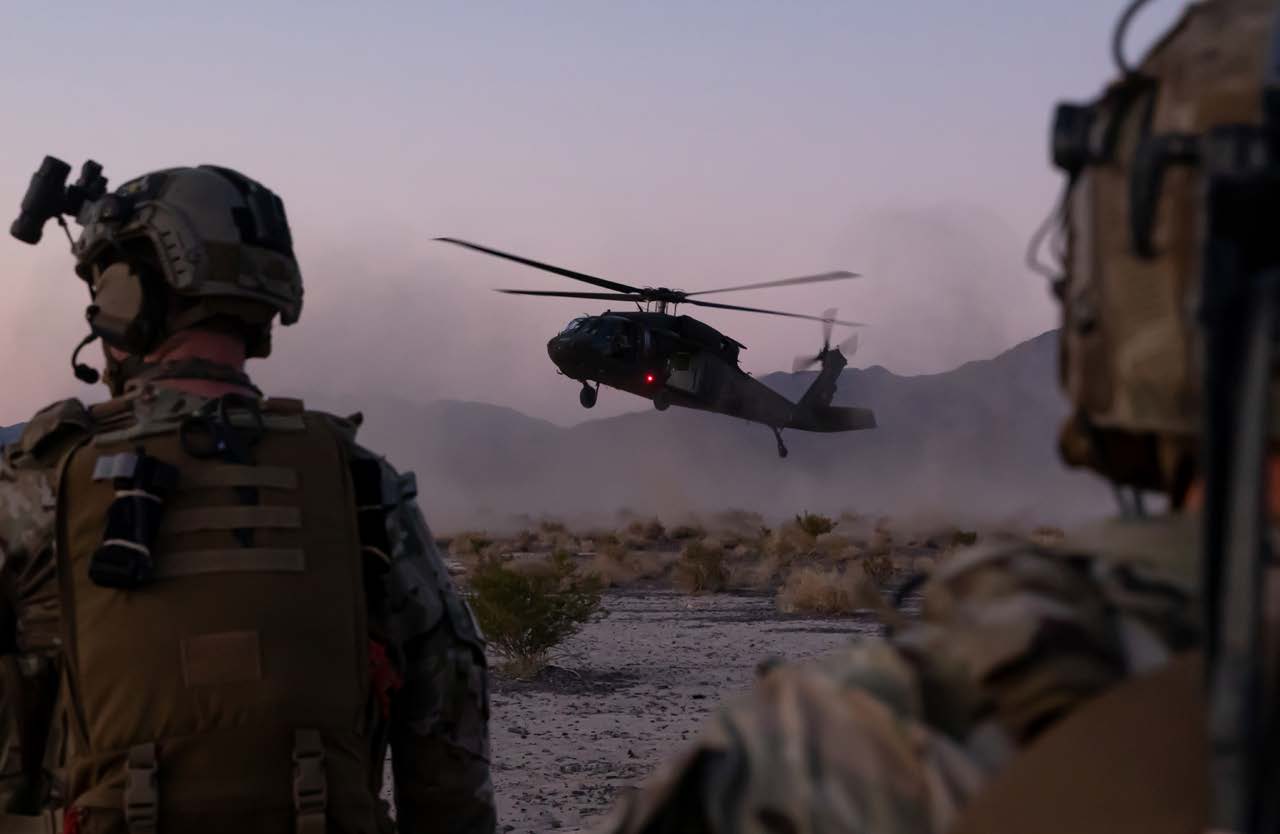 Soldiers at dusk watching a Black Hawk helicopter come in to land across open desert, kicking up dust