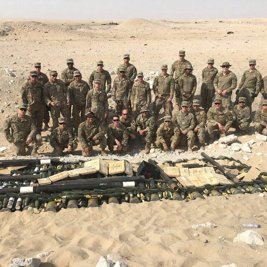 A platoon of U.S. Army soldiers in a desert posed behind a large cache of recovered weapons and ordnance, with Eric kneeling fourth from the left in the front row