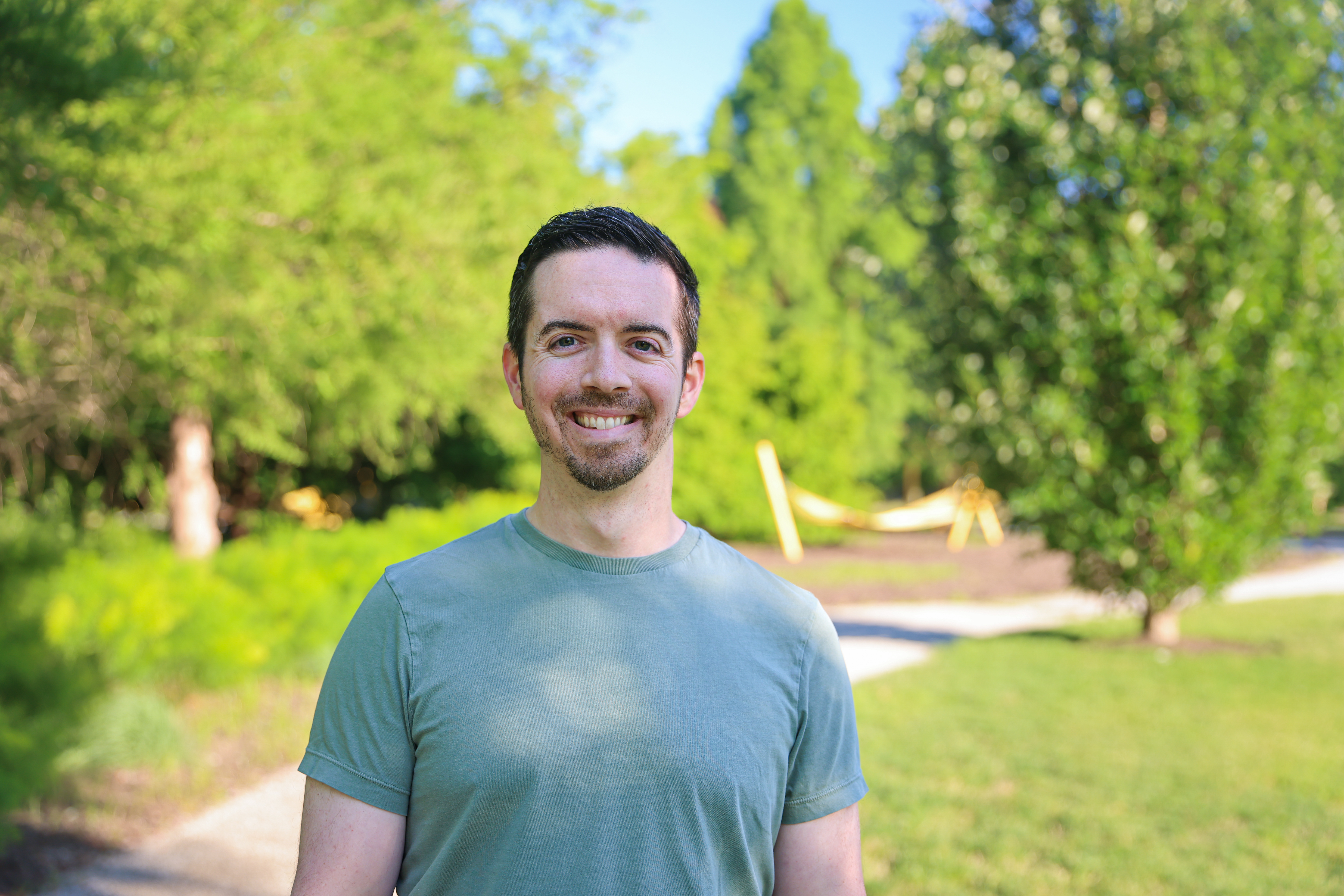Eric D'Aguanno smiling outdoors in a sage-green t-shirt, with sunlit greenery and trees behind him